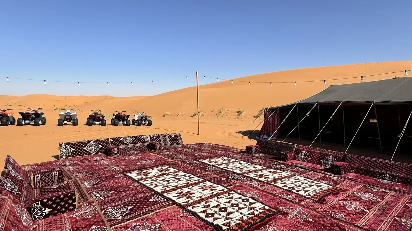 Patterned rugs and low cushions beside a desert tent, with dunes and parked quad bikes beyond.