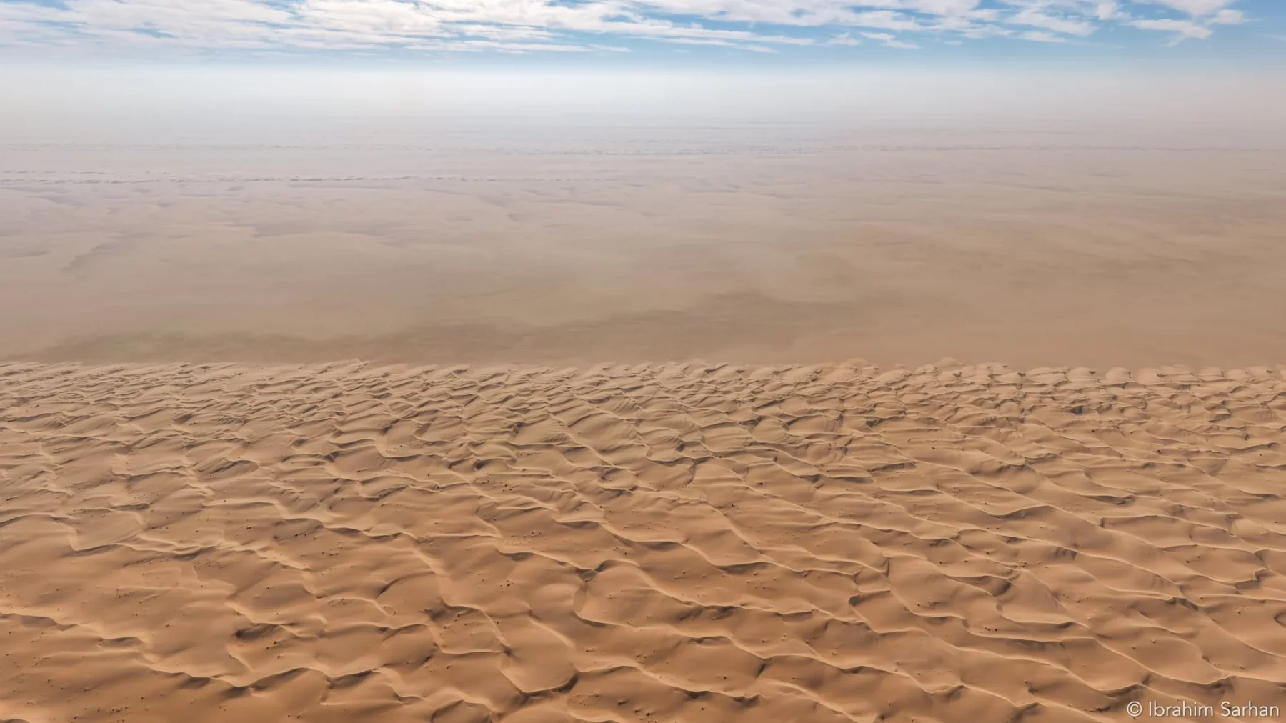 Sand dunes fading into a hazy horizon beneath a blue sky with scattered clouds.