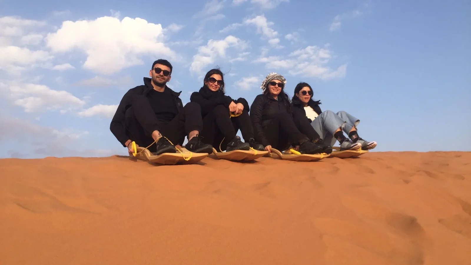 Four visitors seated on sandboards at the top of a dune beneath a cloudy blue sky.