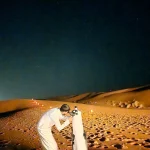 A visitor looking through a telescope on sand dunes at night.