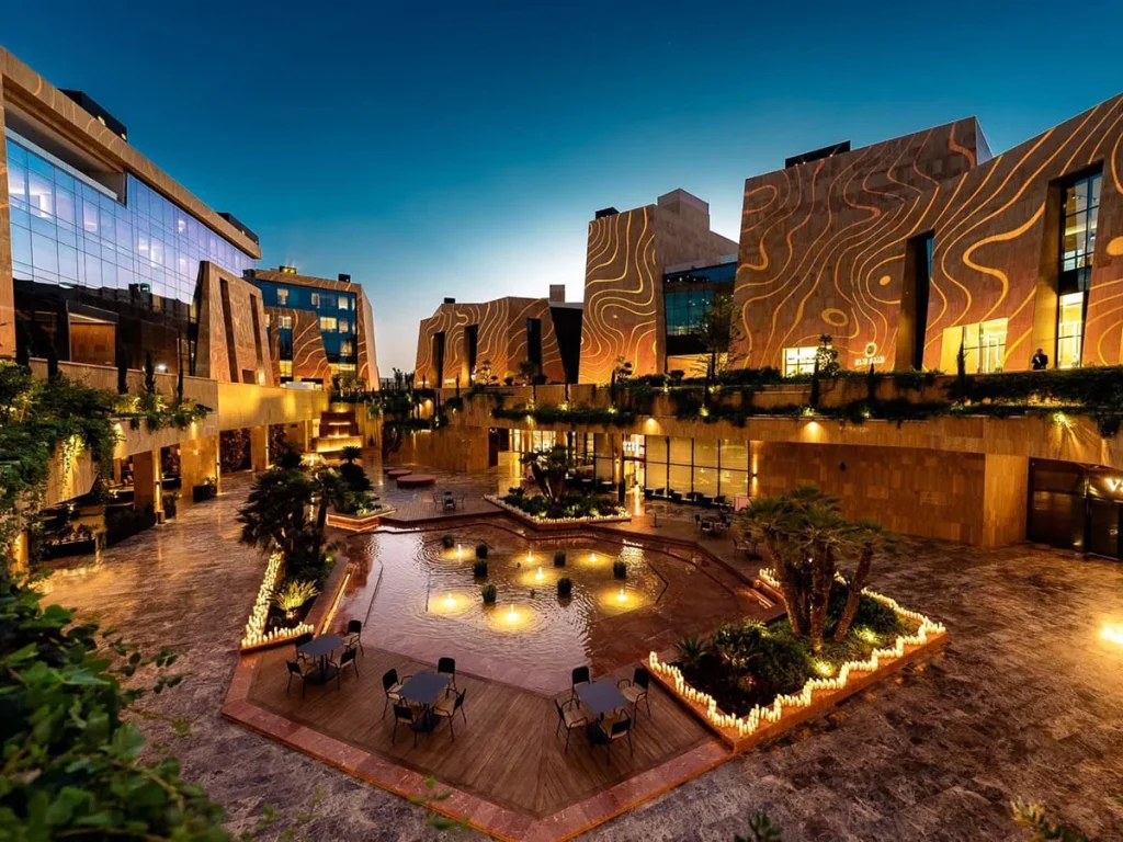Illuminated stone facades surrounding VIA Riyadh’s courtyard, with fountains, palm trees and outdoor tables